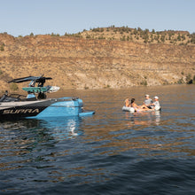 People on an inflatable lounge near a SUPRA boat with a scenic background of cliffs and water.