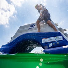 Person standing on a blue and white inflatable boat with Aquaglide Arete 10 branding, on a body of water with a clear sky.