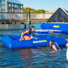 Two people on blue Aquaglide inflatable rafts in a body of water with a dock and house in the background.