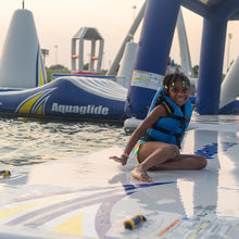 A young girl wearing a blue life vest sits on Aquaglide Speedway 10 at an outdoor water park