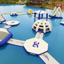 Inflatable water toys and structures on a body of water with a dock and buildings in the background.