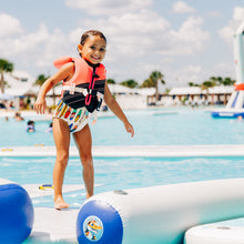 Child in a life jacket standing on an inflatable pool toy with a pool and playground in the background.