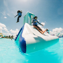 Two people on an inflatable water slide in a pool with a clear blue sky.