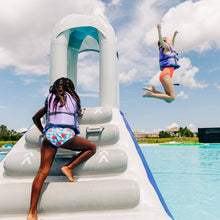 Two children playing on an inflatable water slide by a pool under a blue sky.