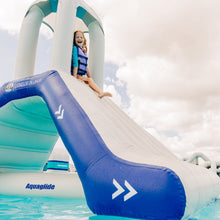 Inflatable water slide with a child on top, branded 'Aquaglide', against a cloudy sky.