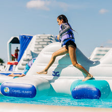 Person playing on an inflatable water slide with 'Camel Humps' branding in a pool.