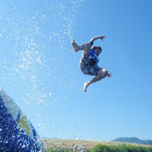 Person jumping off Aquaglide Launch Bag with water splashes against a clear blue sky.