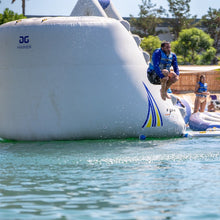 Inflatable water slide with a person jumping off into the water, surrounded by trees and clear sky.