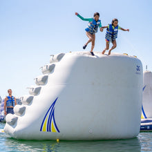 Two children playing on a large inflatable water toy in the water with a clear blue sky.