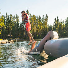A person wearing a red and white striped life vest and orange swim trunks jumps off a Aquaglide Recoil Tramp 17.0 