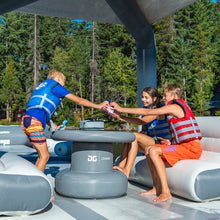 Three children wearing life jackets play on Aquaglide Ohana Lounge in an outdoor lakeside setting with pine trees in the background.