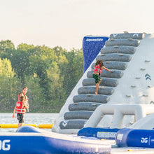 Children playing on an inflatable water slide with Aquaglide branding by a lake.