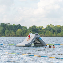 An inflatable water slide on a lake with a child sliding down it, surrounded by water, a tree-lined shore, and a few swimmers in the background.