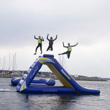 Three people in life vests jumping off an inflatable water slide into a body of water with boats and buildings in the background.