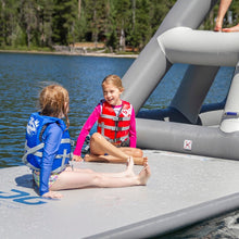 Two children in life vests sitting on an inflatable platform by a lake.