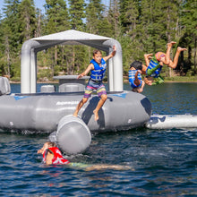 Kids jumping and playing on a gray inflatable water park float on a lake with trees in the background.