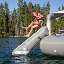 A child wearing a red and white life vest and striped swim trunks jumps from Aquaglide Ricochet Bouncer 12.0 Aquapark into a lake, with a forest shoreline in the background.
