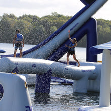 Two people on an inflatable water slide in a lake with trees in the background