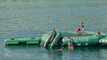 A green inflatable water park platform floating on a lake, with several people jumping, sliding, and swimming around it.