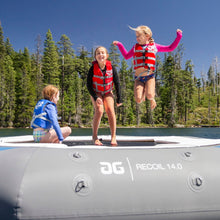 Three children jumping and sitting on a Aquaglide Recoil Tramp 14.0 set against a backdrop of a forested lake under a clear blue sky.
