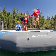 Three children jumping and standing on Aquaglide Recoil Tramp 14.0 on a lake, with a forest and blue sky in the background.