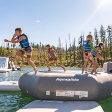 Kids wearing life jackets play on Aquaglide Ricochet Bouncer 12.0 Aquapark on a lake, with trees and a blue sky in the background.