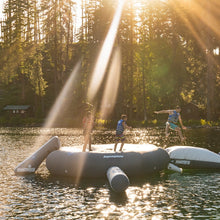 Three people jumping and standing on a large inflatable water platform on a lake, with sun rays shining through trees in the background.
