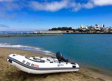 Island Hopper Shark 9.8′ Inflatable Fishing Boat | Aluminum Floor Dinghy  on a sandy beach next to blue water, with a city skyline in the background under a blue sky.