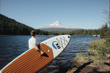 A person stands at the edge of a lake holding Island Hopper Inflatable Sport Board AIRBO 10.6′ ISUP & Kayak
