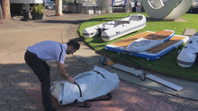 A man in a white shirt and black pants is packing a white inflatable dinghy bag outdoors on a paved walkway. Several inflatable boats and boards are arranged on grass in the background.