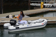 A woman sits on a Island Hopper Shark 9.8′ Inflatable Fishing Boat | Aluminum Floor Dinghy with a black Mercury outboard motor, docked at a harbor. 