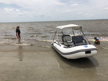 A white inflatable boat with a white canopy and black seating is parked on a sandy beach in shallow water, with two people standing in the ocean nearby under a cloudy sky.
