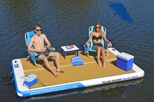 Two people on an inflatable dock with chairs, cooler, and umbrella in a body of water.